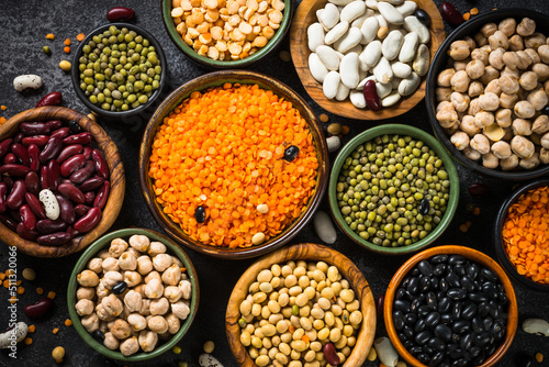 Legumes, lentils, chikpea and beans assortment in different bowls on black stone table. Top view.