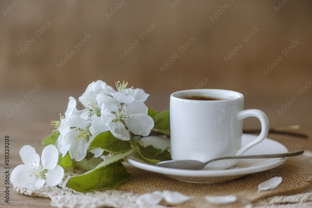 Coffee in a white cup on a white saucer with a teaspoon and a branch of a flowering apple tree. Morning breakfast with coffee. Coffee in a white cup on a white saucer with a teaspoon and a branch of a flowering apple tree. Morning breakfast with coffee.