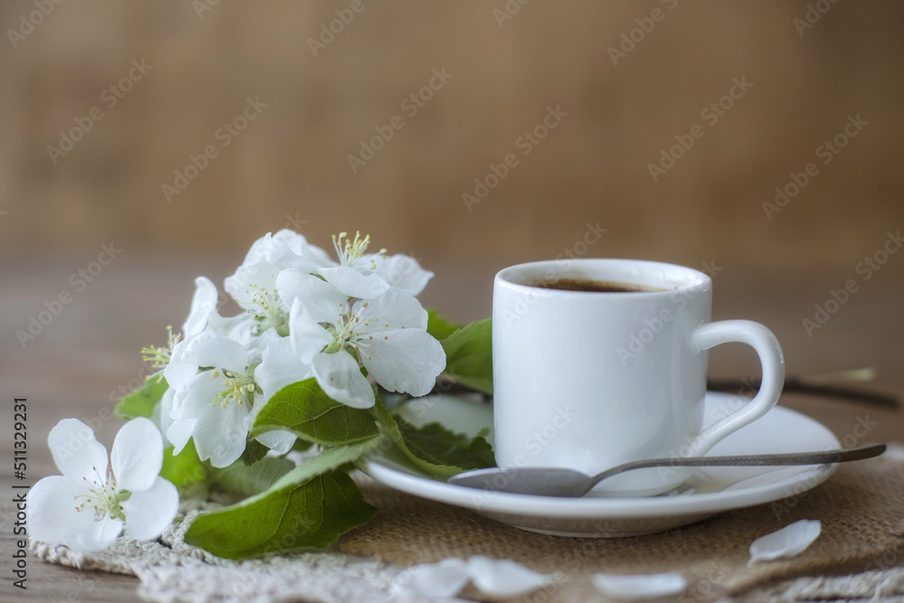 Coffee in a white cup on a white saucer with a teaspoon and a branch of a flowering apple tree. Morning breakfast with coffee. Coffee in a white cup on a white saucer with a teaspoon and a branch of a flowering apple tree. Morning breakfast with coffee.