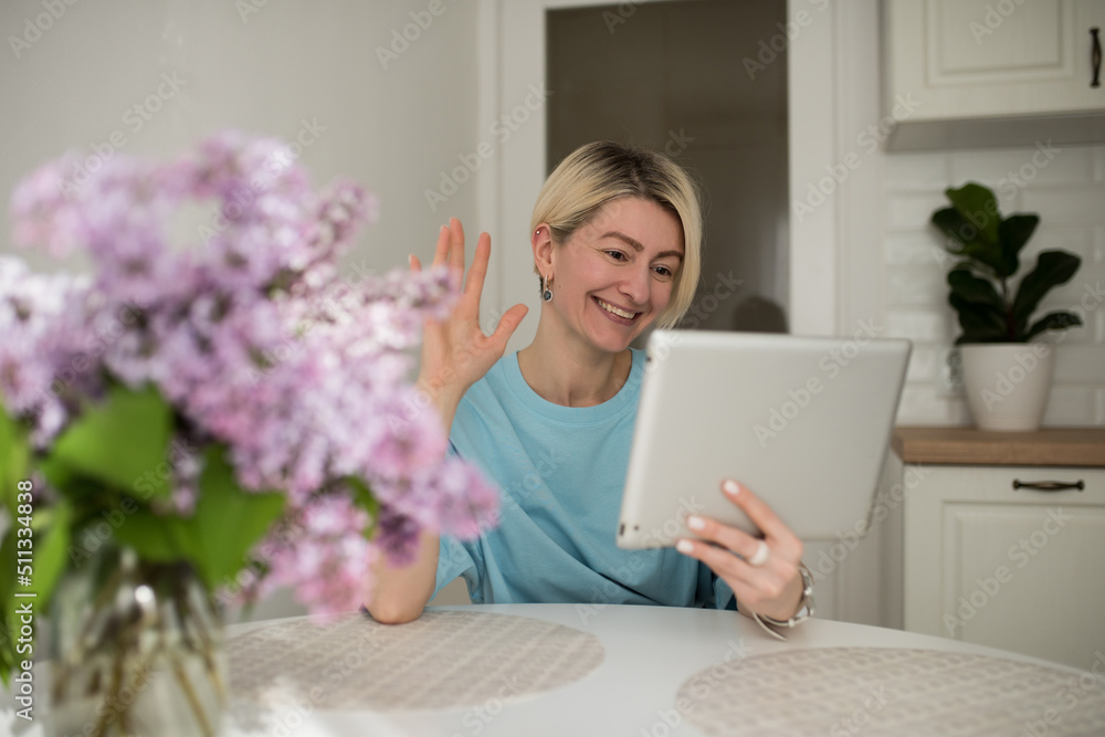 Happy woman with blond hair using digital tablet to make video calls to parents and friends, smiling girl sitting at home in the kitchen cheerfully greets online using a computer webcam Happy woman with blond hair using digital tablet to make video calls to parents and friends, smiling girl sitting at home in the kitchen cheerfully greets online using a computer webcam