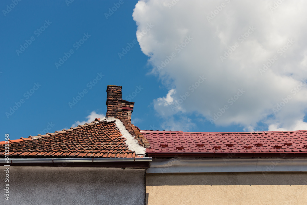 Old chimney made of bricks on a roof with ceramic tiles with a cloud above the home next to a house with new metallic rooftop. Construction backdrop