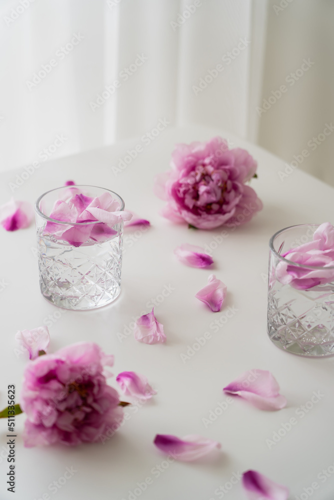 fresh peonies and petals near glasses with gin tonic on white tabletop and grey background.