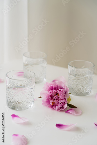 pink peony and floral petals near glasses with fresh water on grey background.