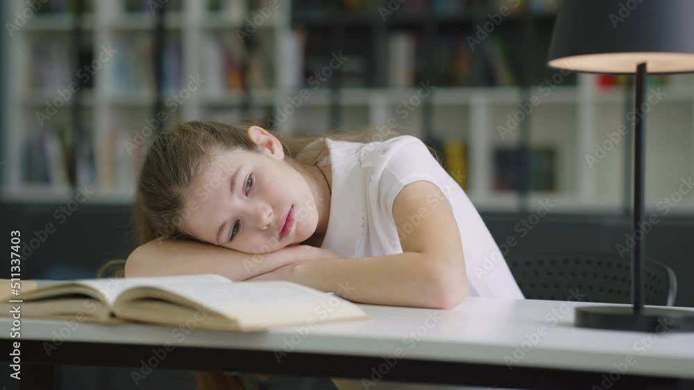 Sad girl lying on book and looking to side, pupil feeling tired or depressed, bookshelves on blurred background. Concept of education Sad girl lying on book and looking to side, pupil feeling tired or depressed, bookshelves on blurred background. Concept of education