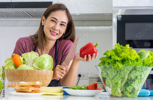  Asian woman stands at the kitchen counter holding a bell pepper and uses a vegetable knife to make a healthy salad with healthy nutrition. photo