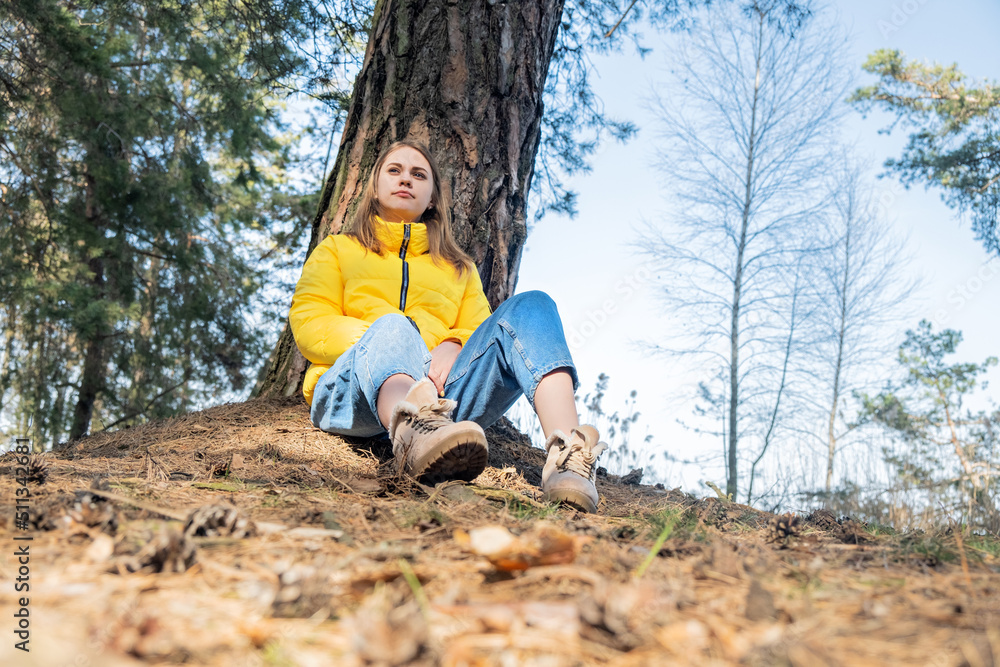 Portrait of a happy smiling young girl sitting in the woods under a tree. Mental health. The woman wears a bright yellow jacket. Time for yourself, reboot, meditation. Portrait of a happy smiling young girl sitting in the woods under a tree. Mental health. The woman wears a bright yellow jacket. Time for yourself, reboot, meditation.