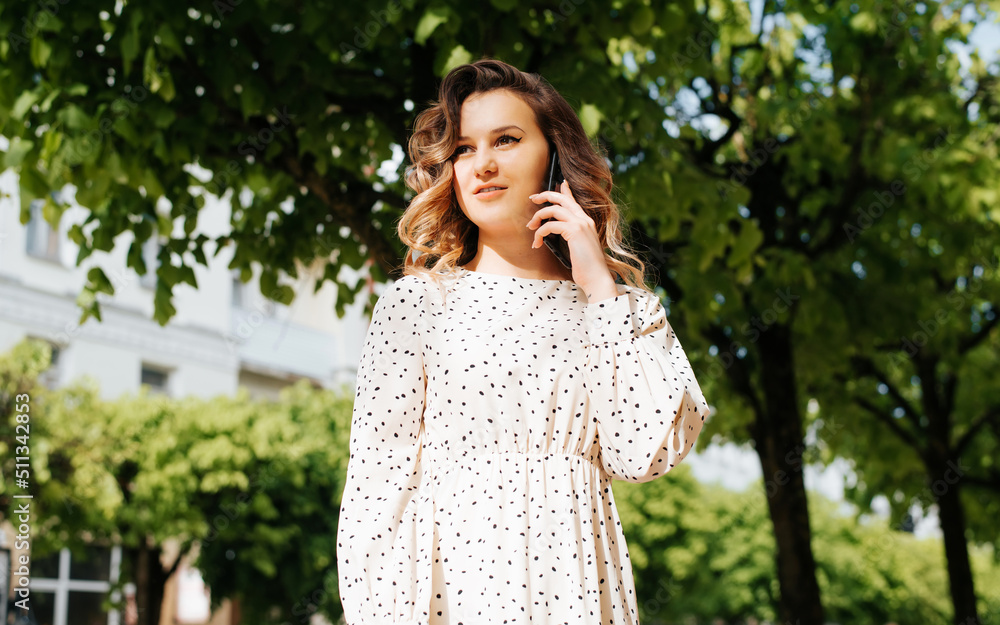 Portrait of young woman calling on smartphone and looking away while standing outdoors in park on sunny day. Curly pretty lady in white summer dress using mobile phone Portrait of young woman calling on smartphone and looking away while standing outdoors in park on sunny day. Curly pretty lady in white summer dress using mobile phone