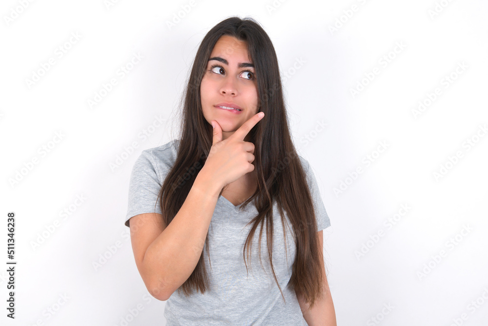 Young beautiful brunette woman wearing grey T-shirt over white wall Thinking worried about a question, concerned and nervous with hand on chin. Young beautiful brunette woman wearing grey T-shirt over white wall Thinking worried about a question, concerned and nervous with hand on chin.