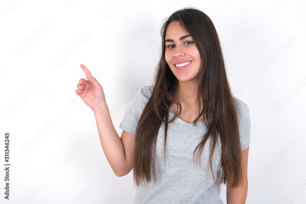 Young beautiful brunette woman wearing grey T-shirt over white wall looking at camera indicating finger empty space sales Young beautiful brunette woman wearing grey T-shirt over white wall looking at camera indicating finger empty space sales