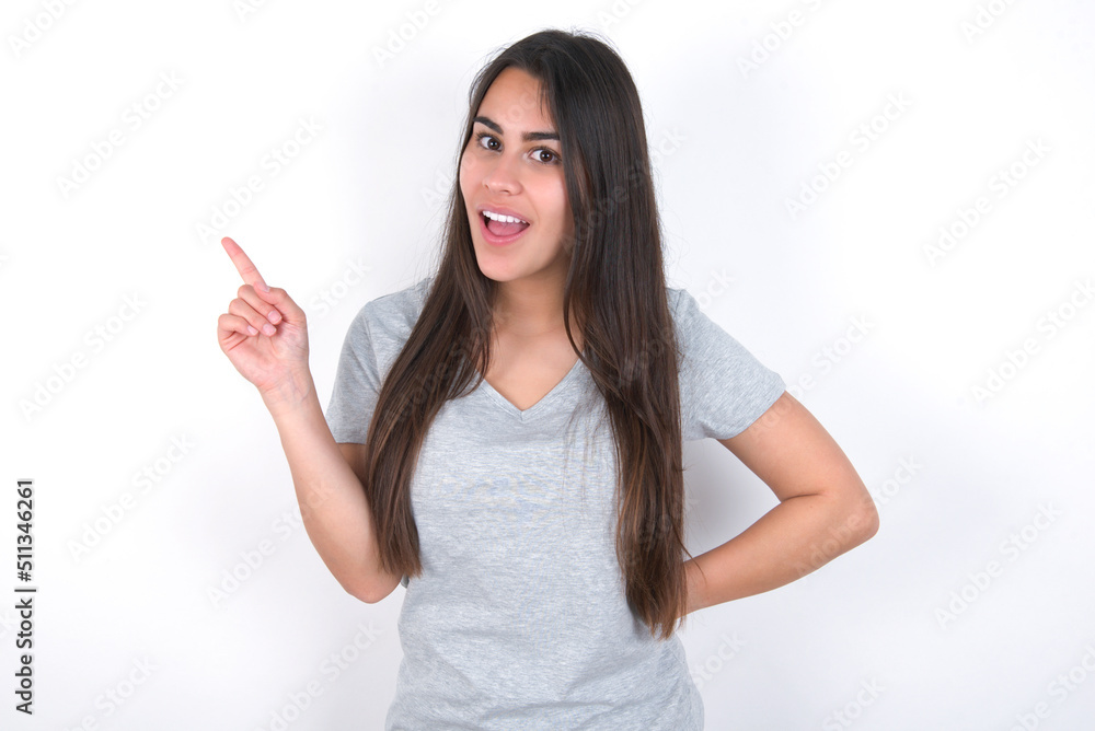 Young beautiful brunette woman wearing grey T-shirt over white wall holding finger up having idea and posing Young beautiful brunette woman wearing grey T-shirt over white wall holding finger up having idea and posing