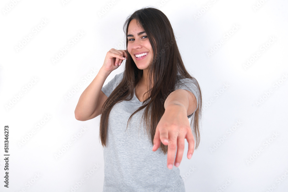 Young beautiful brunette woman wearing grey T-shirt over white wall smiling cheerfully and pointing to camera while making a call you later gesture, talking on phone Young beautiful brunette woman wearing grey T-shirt over white wall smiling cheerfully and pointing to camera while making a call you later gesture, talking on phone