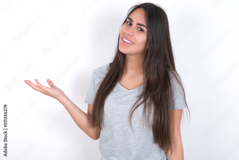 Young beautiful brunette woman wearing grey T-shirt over white wall smiling cheerful presenting and pointing with palm of hand looking at the camera. Young beautiful brunette woman wearing grey T-shirt over white wall smiling cheerful presenting and pointing with palm of hand looking at the camera.