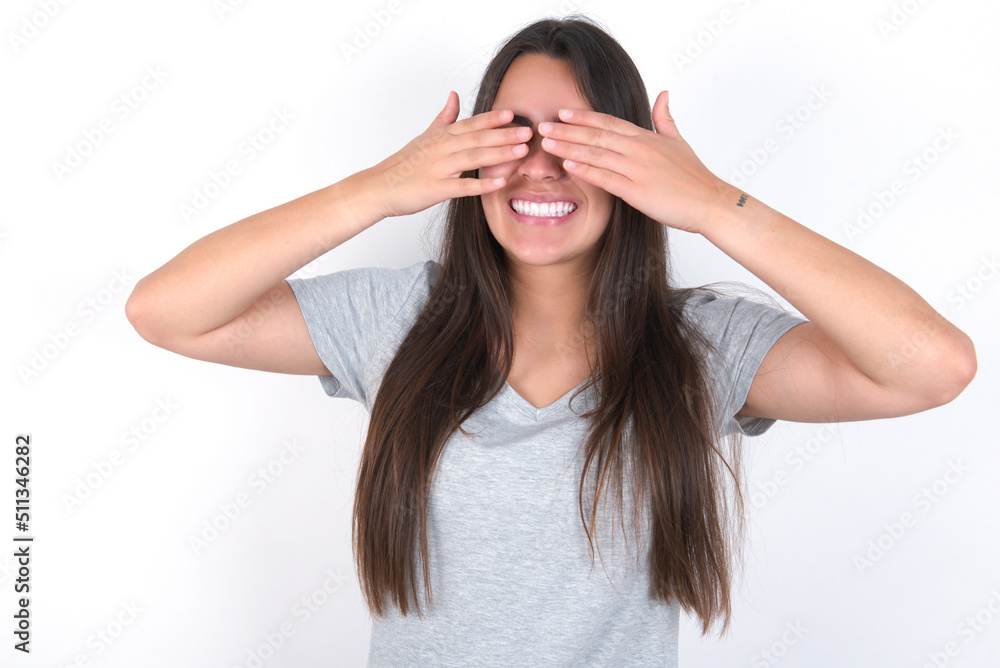 Young beautiful brunette woman wearing grey T-shirt over white wall covering eyes with hands smiling cheerful and funny. Blind concept. Young beautiful brunette woman wearing grey T-shirt over white wall covering eyes with hands smiling cheerful and funny. Blind concept.