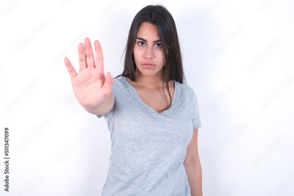 young beautiful brunette woman wearing grey t-shirt over white wall doing stop sing with palm of the hand. Warning expression with negative and serious gesture on the face. young beautiful brunette woman wearing grey t-shirt over white wall doing stop sing with palm of the hand. Warning expression with negative and serious gesture on the face.