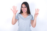 young beautiful brunette woman wearing grey t-shirt over white wall showing and pointing up with fingers number eight while smiling confident and happy.