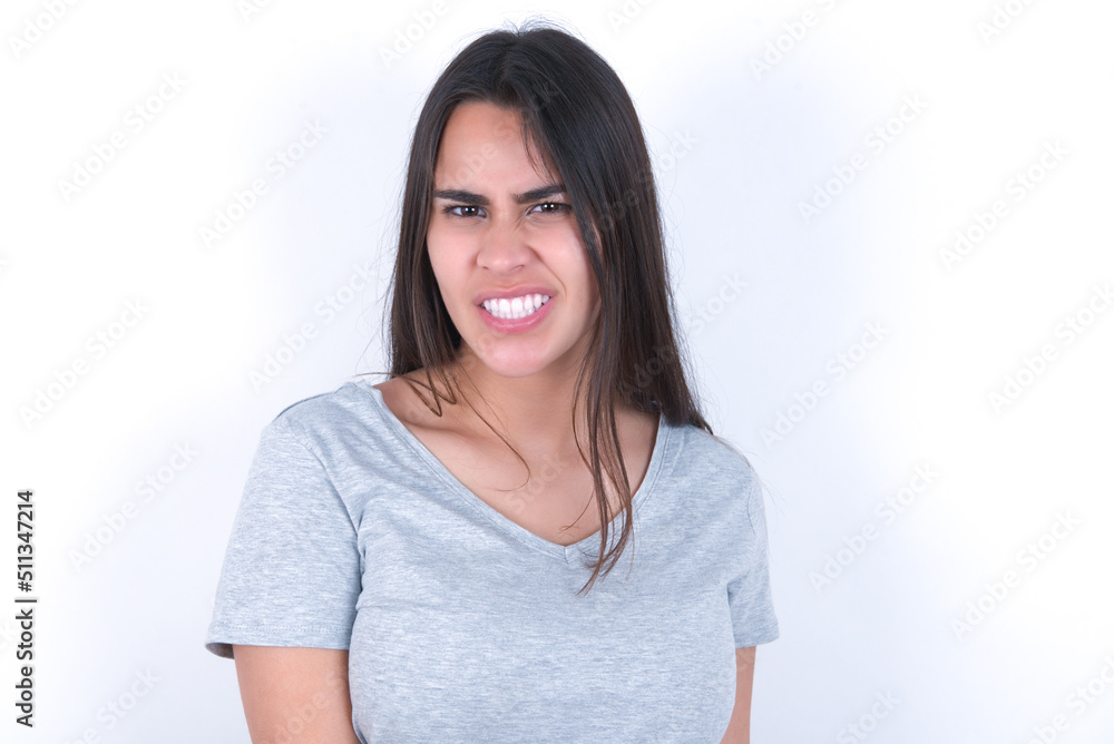 Fototapeta premium Young beautiful brunette woman wearing gray t-shirt over white wall keeps teeth clenched, frowns face in dissatisfaction, irritated because of much duties.