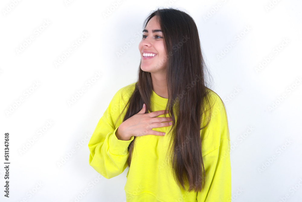 Joyful Young beautiful brunette woman wearing yellow hoodie over white wall expresses positive emotions recalls something funny keeps hand on chest and giggles happily. Joyful Young beautiful brunette woman wearing yellow hoodie over white wall expresses positive emotions recalls something funny keeps hand on chest and giggles happily.