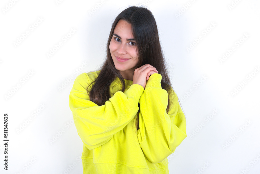 Charming serious Young beautiful brunette woman wearing yellow hoodie over white wall keeps hands near face smiles tenderly at camera Charming serious Young beautiful brunette woman wearing yellow hoodie over white wall keeps hands near face smiles tenderly at camera