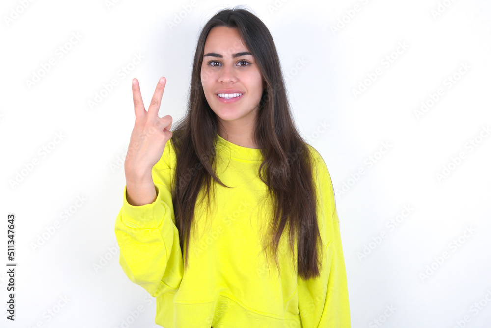 Young beautiful brunette woman wearing yellow hoodie over white wall smiling and looking friendly, showing number two or second with hand forward, counting down Young beautiful brunette woman wearing yellow hoodie over white wall smiling and looking friendly, showing number two or second with hand forward, counting down