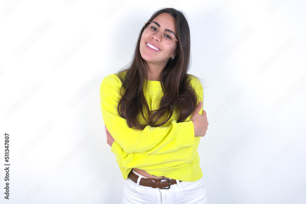 Charming pleased Young beautiful brunette woman wearing yellow hoodie over white wall embraces own body, pleasantly feels comfortable poses. Tenderness and self esteem concept Charming pleased Young beautiful brunette woman wearing yellow hoodie over white wall embraces own body, pleasantly feels comfortable poses. Tenderness and self esteem concept