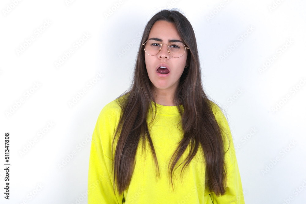 Young beautiful brunette woman wearing yellow hoodie over white wall yawns with opened mouth stands. Daily morning routine Young beautiful brunette woman wearing yellow hoodie over white wall yawns with opened mouth stands. Daily morning routine