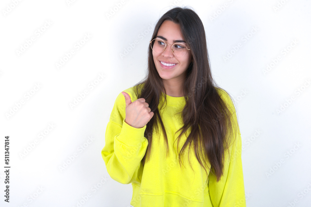 Young beautiful brunette woman wearing yellow hoodie over white wall points away and gives advice demonstrates advertisement Young beautiful brunette woman wearing yellow hoodie over white wall points away and gives advice demonstrates advertisement