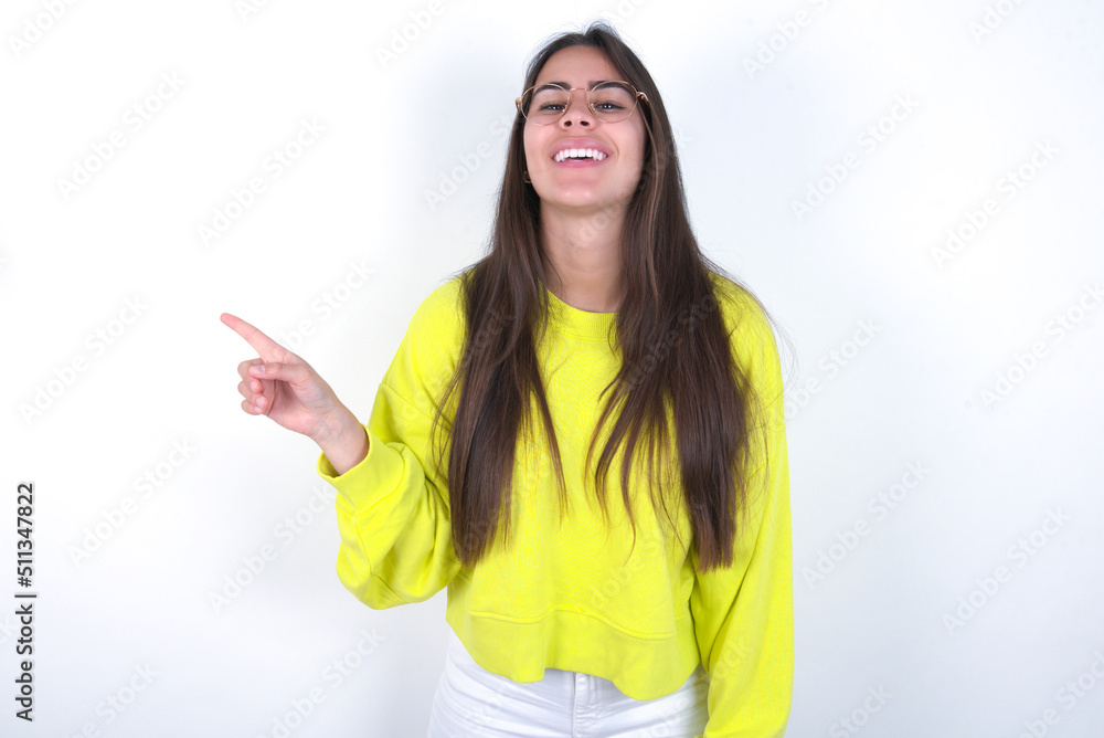Young beautiful brunette woman wearing yellow hoodie over white wall laughs happily points away on blank space demonstrates shopping discount offer, excited by good news or unexpected sale. Young beautiful brunette woman wearing yellow hoodie over white wall laughs happily points away on blank space demonstrates shopping discount offer, excited by good news or unexpected sale.