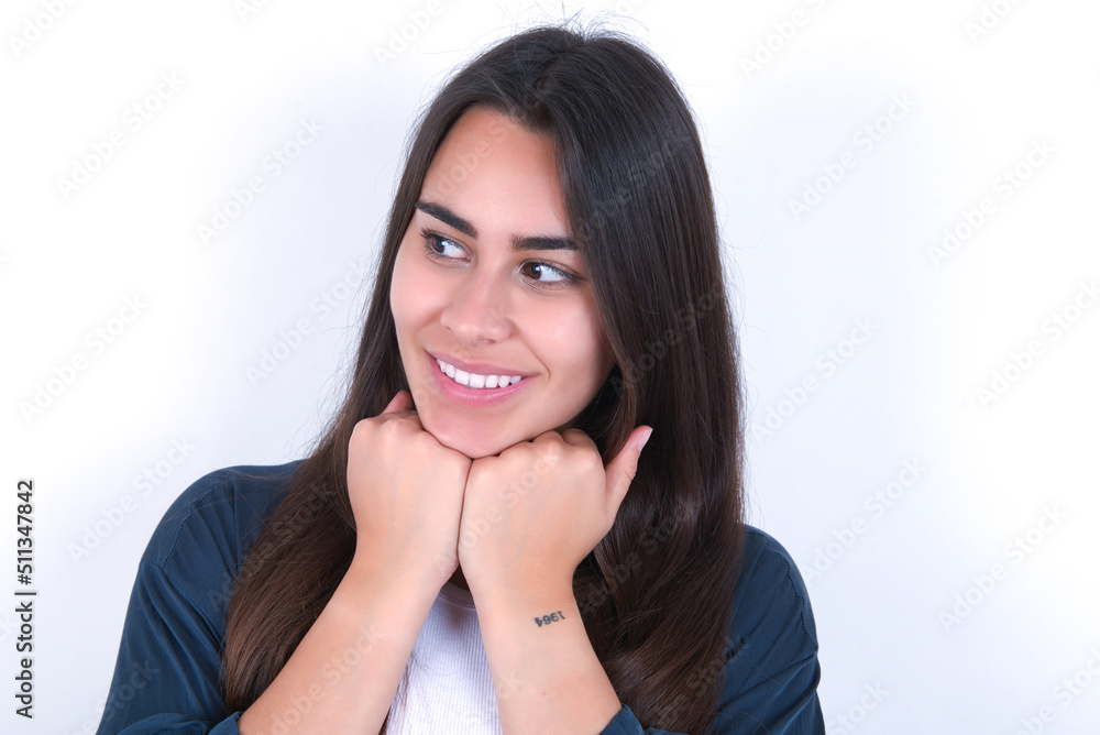 Young beautiful brunette woman wearing blue jacket over white wall, holds hands under chin, glad to hear heartwarming words from stranger Young beautiful brunette woman wearing blue jacket over white wall, holds hands under chin, glad to hear heartwarming words from stranger