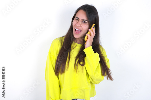 Funny Young beautiful brunette woman wearing yellow hoodie over white wall laughs happily, has phone conversation, being amused by friend, closes eyes.