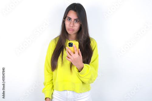 Upset dissatisfied Young beautiful brunette woman wearing yellow hoodie over white wall uses mobile software application and surfs information in internet, holds modern mobile hand