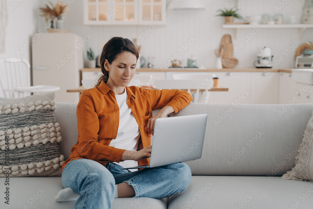 Naklejka premium Happy young woman with laptop is sitting on couch at home. Remote work or study from home.