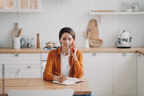 Young woman is call center assistant. Girl is sitting at table at kitchen and taking notes.