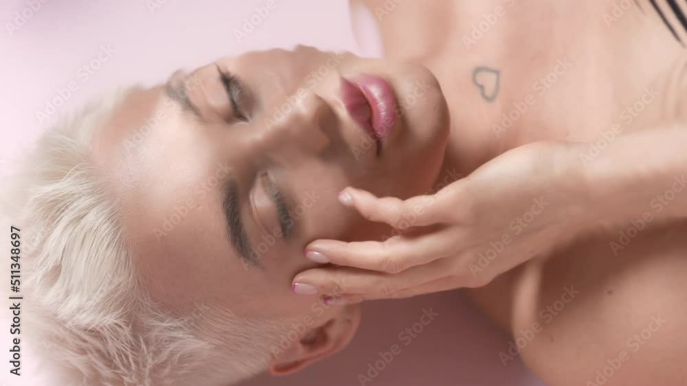 Good-looking young slim Caucasian woman with short light hair turns her head to the camera, opens her eyes and strokes her face through rippling water on pale pink background | Moisturizing commercial Good-looking young slim Caucasian woman with short light hair turns her head to the camera, opens her eyes and strokes her face through rippling water on pale pink background | Moisturizing commercial