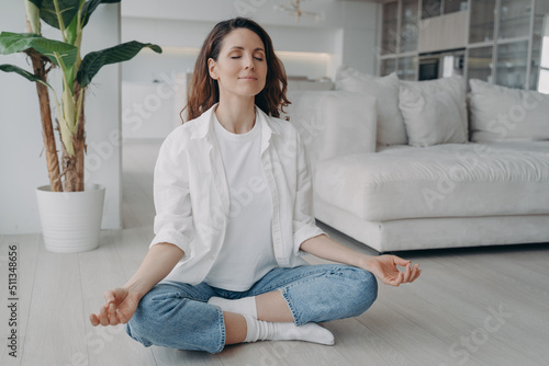 Caucasian woman is sitting in lotus asana at home in morning. Scandinavian interior of living room. photo