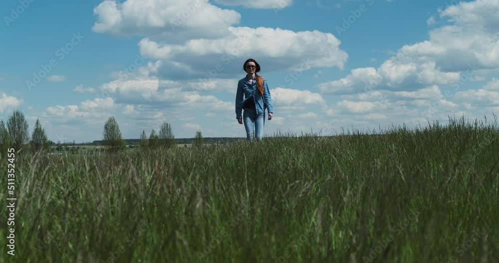 Young woman in denim is walking towards the green tall grass. White clouds, blue sky, spring summer. Concept of return, go to your goal, confidence, countryside. Wide shot, daytime, sunny. Young woman in denim is walking towards the green tall grass. White clouds, blue sky, spring summer. Concept of return, go to your goal, confidence, countryside. Wide shot, daytime, sunny.