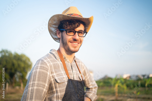 Portrait of young Caucasian male farmer in apron  glasses  and straw hat turning to smiling at camera while standing in farmland with a blurred banana plantation and clear blue sky in the background.