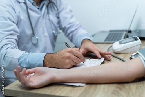 Focus on Caucasian male doctor's hand in white gown taking notes on clipboard while measured Asian patient's pressure and pulse rate by using digital blood pressure monitor on examination room table.