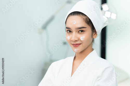 Beautiful Asian woman in bathrobe and towel on head with facial treatment cream applied on her face, smiling looking at camera while doing a beauty treatments procedures in a clinic treatment room.