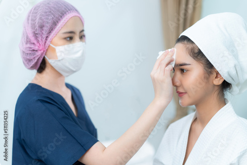 Asian female beautician in blue clothes, medical hat, and face mask using a cotton pad wiping on beautiful Asian female client's face who smiling sitting on bed in a beauty clinic's treatment room.