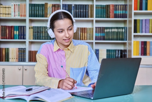 High school student girl in headphones studying in school library using books and laptop