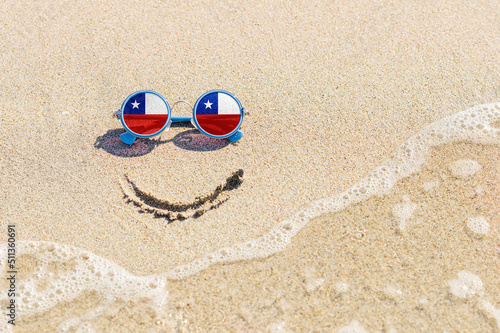 A painted smile on the beach and sunglasses with the flag of the Chile. The concept of a positive holiday in the resort of the Chile.
