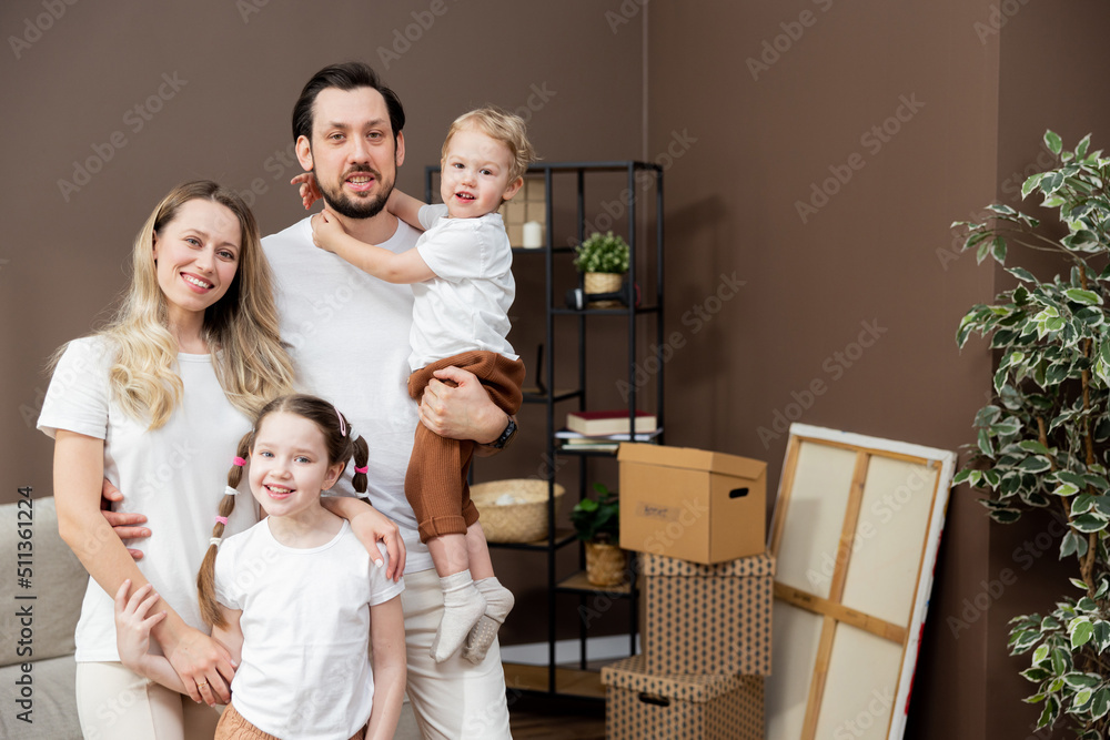 Portrait of happy young Caucasian family with kids standing in new home, holding cardboard boxes, smiling and looking at camera. Real estate, buying property, moving house or apartment concept. Portrait of happy young Caucasian family with kids standing in new home, holding cardboard boxes, smiling and looking at camera. Real estate, buying property, moving house or apartment concept.