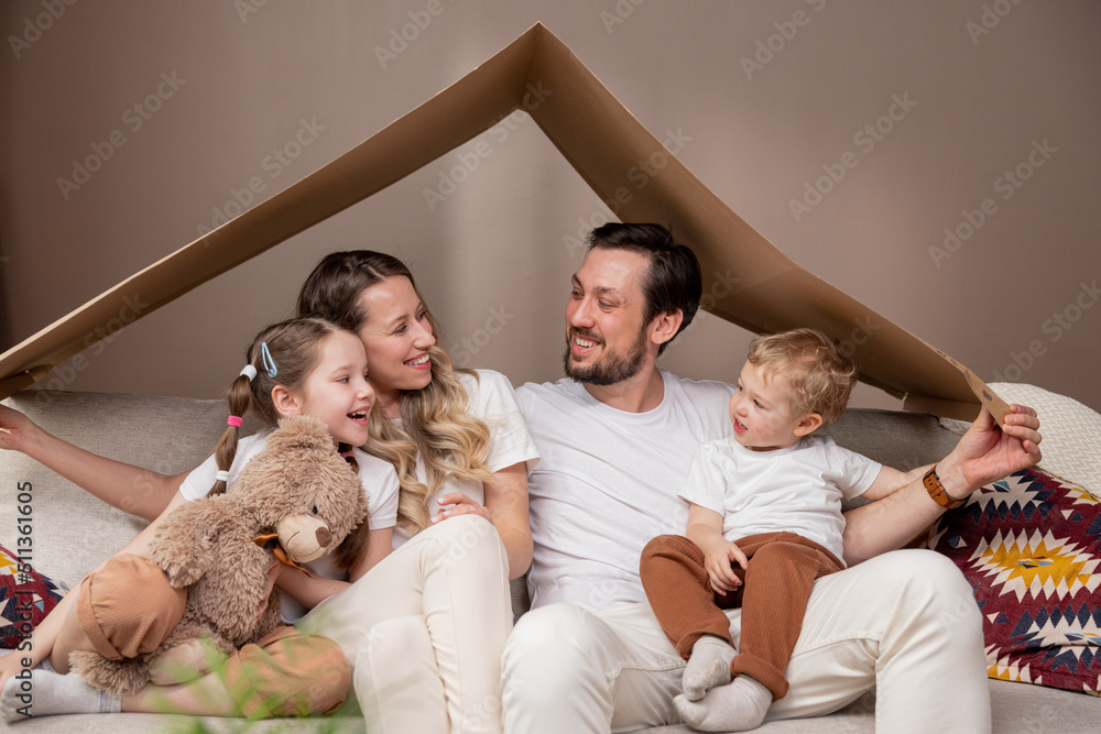 Family complete parents with two children sitting on floor relaxing after moving to new apartment holding piece of cardboard left over from boxes as roof over their heads smiling to camera each other.