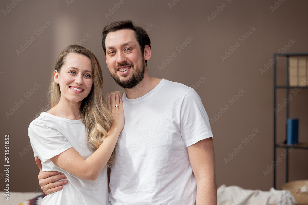 Smiling woman hugging husband from the side on the living room wall background. Man proudly embraces his beautiful young wife. Freshly married couple love family. Smiling woman hugging husband from the side on the living room wall background. Man proudly embraces his beautiful young wife. Freshly married couple love family.