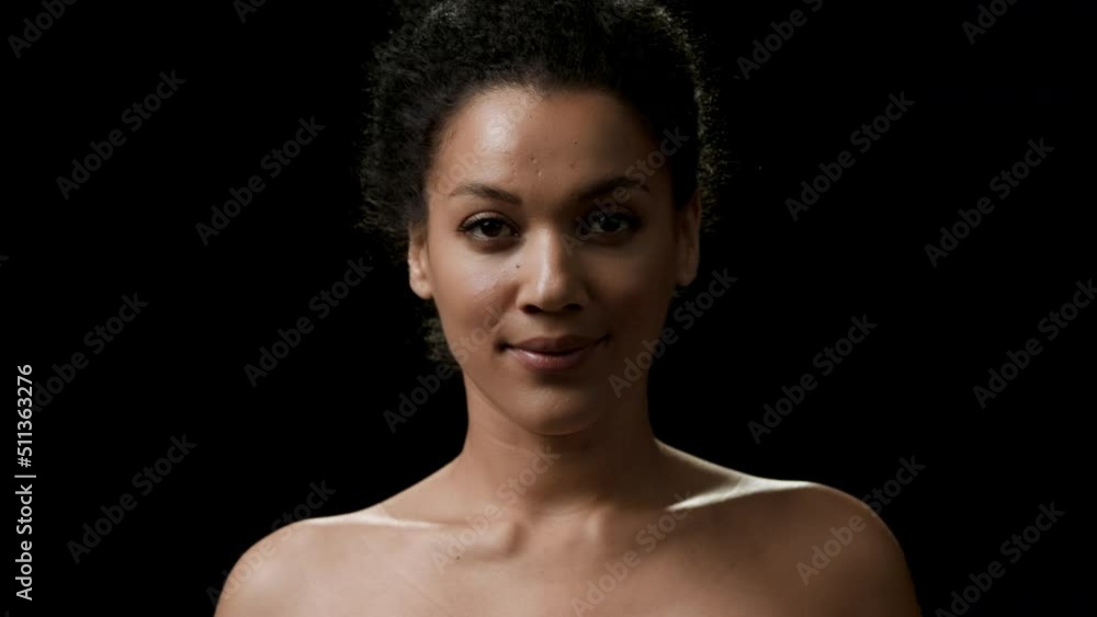 Black-haired young slim African American good-looking woman with curly hair in bun turns to the camera smiling for it on black background | Body care concept Black-haired young slim African American good-looking woman with curly hair in bun turns to the camera smiling for it on black background | Body care concept