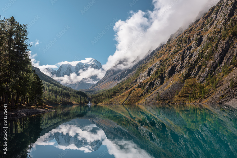 Obraz premium Scenic landscape with large snow mountains in low clouds reflected in turquoise mountain lake mirror in morning sunlight. Alpine lake in autumn valley with view to sunlit high snowy mountain range.