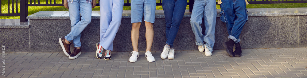 Group of people in casual clothes outdoors. Several young men and women in trendy jeans, shorts, pants and sneakers standing by fence on city street. Cropped shot, human legs. Fashion concept banner