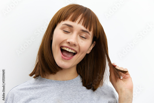 a beautiful, happy woman stands on a white background in light clothes with straight, red hair and pulling one strand smiles broadly with her eyes closed