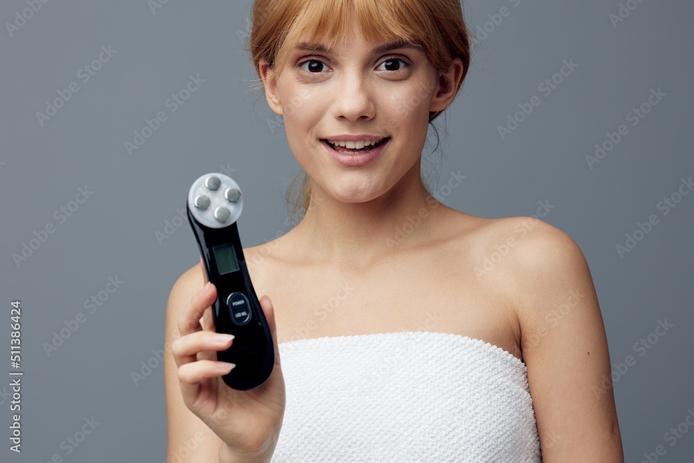 a woman with perfect skin is standing on a gray background wrapped in a towel and looking at the camera holding an electric massager smiling pleasantly. Close horizontal photo