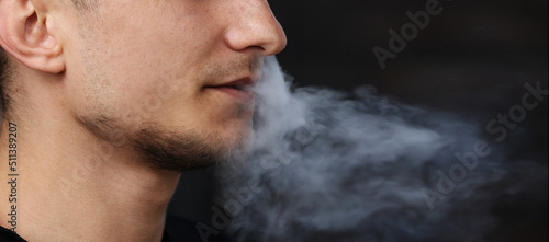close up of a man with an electronic cigarette. man smokes an electronic cigarette in a natural background. hipster smokes an e-cigarette, breathes out streams of smoke and vaping outdoors copy space